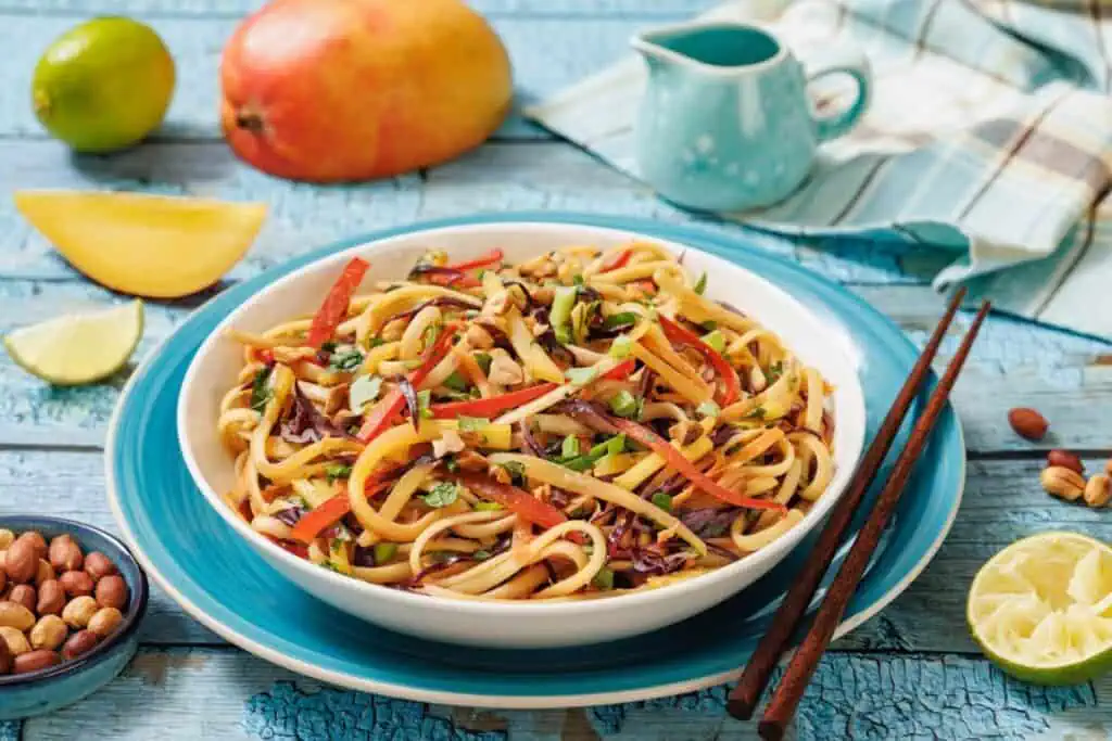 A bowl of noodle salad with vegetables, garnished with peanuts and herbs, sits on a blue plate with chopsticks, surrounded by a mango, lime, peanuts, and a small pitcher.
