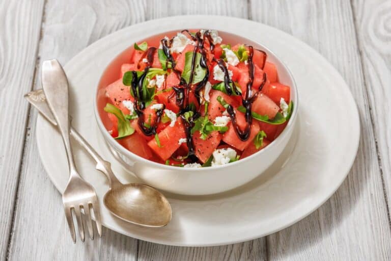 A bowl of watermelon salad with feta cheese, arugula, and balsamic glaze, placed on a white plate with a fork and spoon on the side.