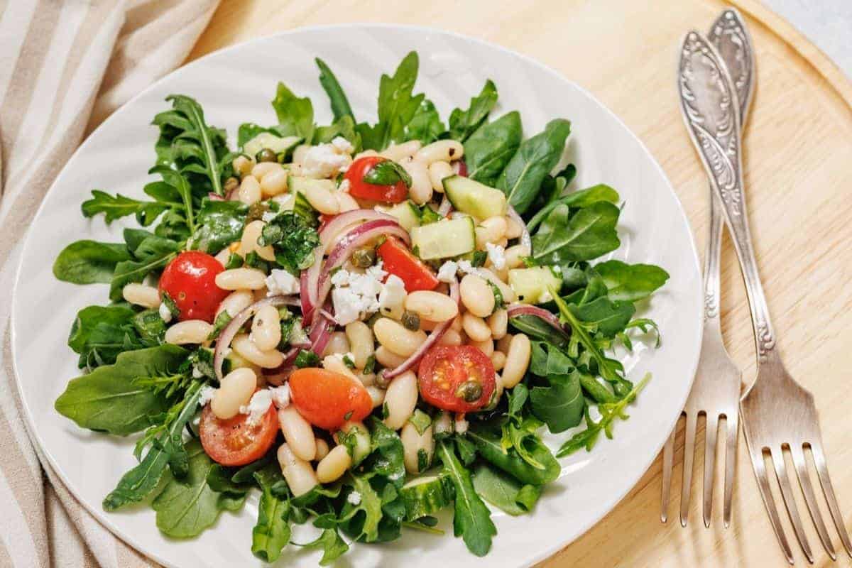 A fresh White Bean Salad on a white plate featuring arugula, white beans, cherry tomatoes, sliced red onions, avocado, and cheese, placed on a wooden surface with two forks beside it.