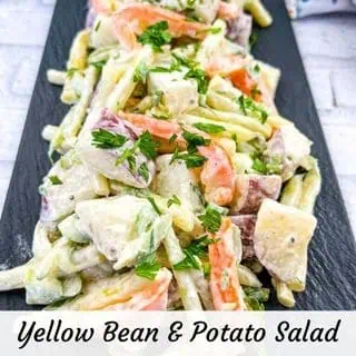 A close-up of yellow bean and potato salad with carrots, cubed potatoes, and fresh parsley on a black serving tray. Text at the bottom reads “Yellow Bean & Potato Salad.”.