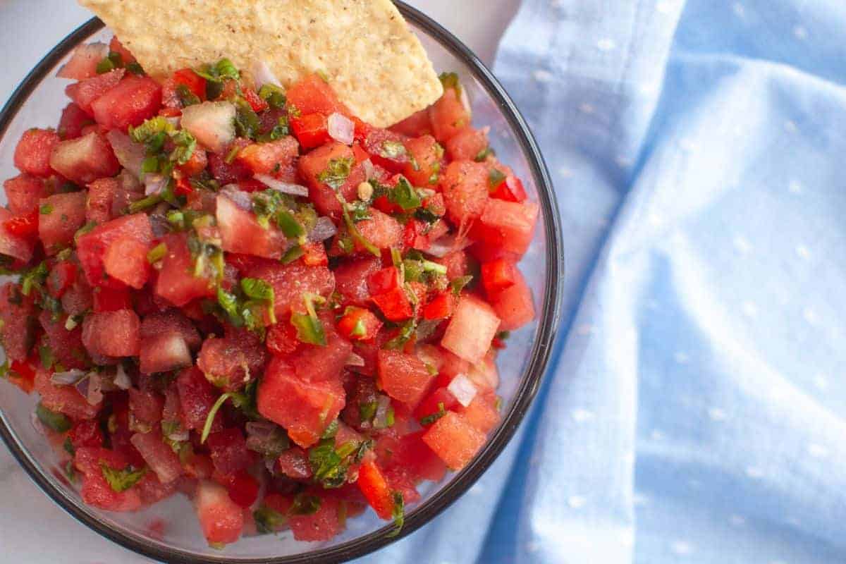 A bowl of watermelon salsa with chopped herbs and vegetables, served with a tortilla chip, next to a light blue cloth.