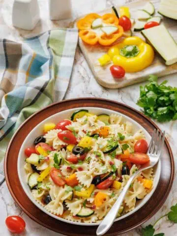 A bowl of bowtie pasta salad with cherry tomatoes, bell peppers, zucchini, olives, and parsley sits on a plate next to a fork, with fresh vegetables and herbs in the background.