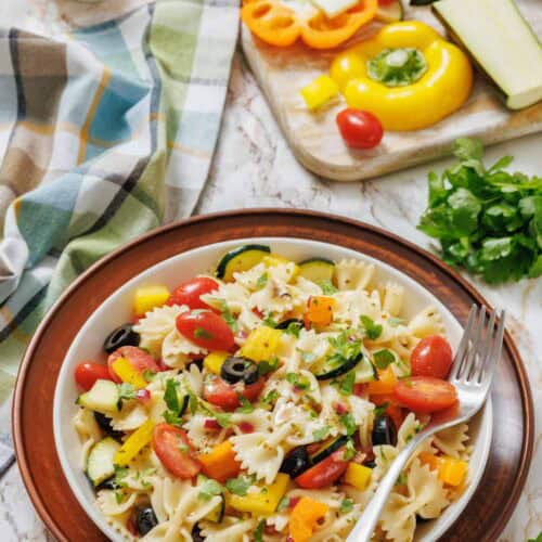 A bowl of bowtie pasta salad with cherry tomatoes, bell peppers, zucchini, olives, and parsley sits on a plate next to a fork, with fresh vegetables and herbs in the background.