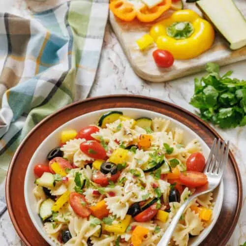 A bowl of bowtie pasta salad with cherry tomatoes, bell peppers, zucchini, olives, and parsley sits on a plate next to a fork, with fresh vegetables and herbs in the background.