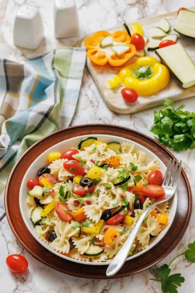 A bowl of bowtie pasta salad with cherry tomatoes, bell peppers, zucchini, olives, and parsley sits on a plate next to a fork, with fresh vegetables and herbs in the background.