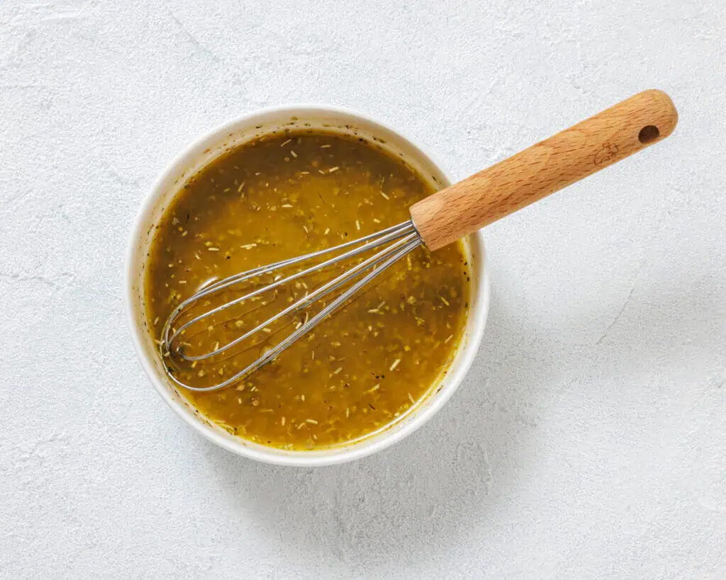 A bowl of yellow-green vinaigrette dressing with visible herbs, with a metal whisk resting inside.
