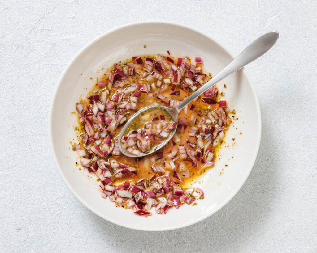 A white bowl with chopped red onions, oil, herbs, and vinegar being mixed with a spoon on a light surface.
