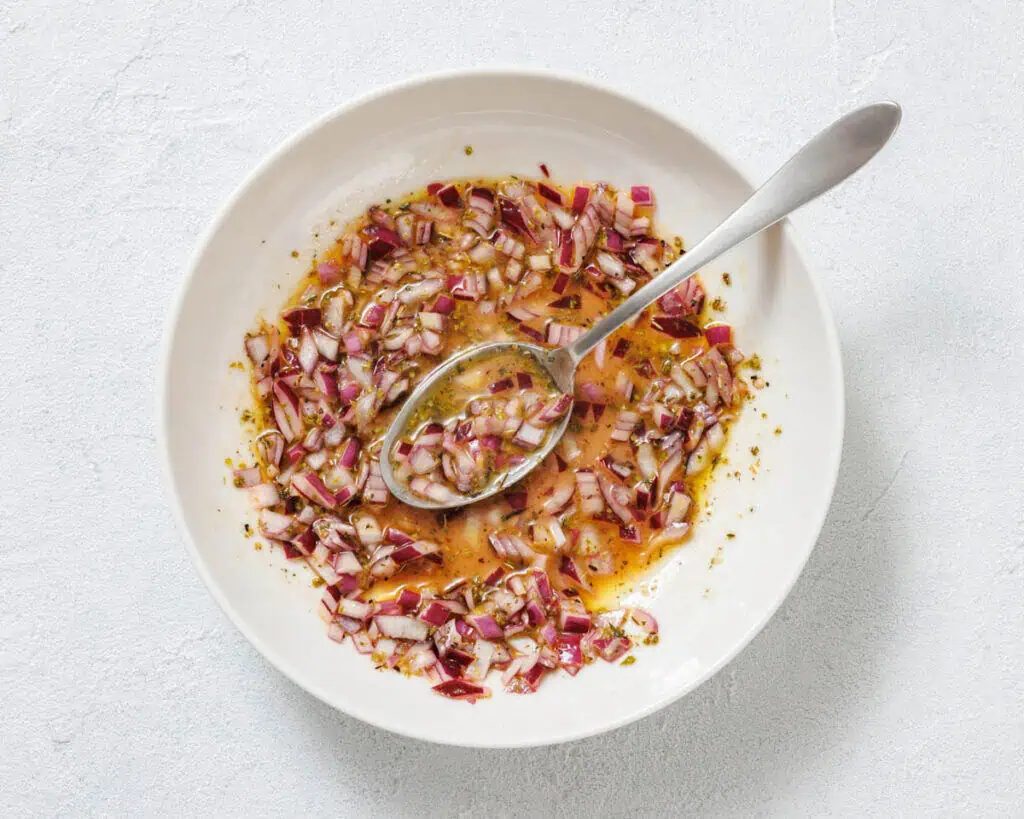 A white bowl with chopped red onions, oil, herbs, and vinegar being mixed with a spoon on a light surface.