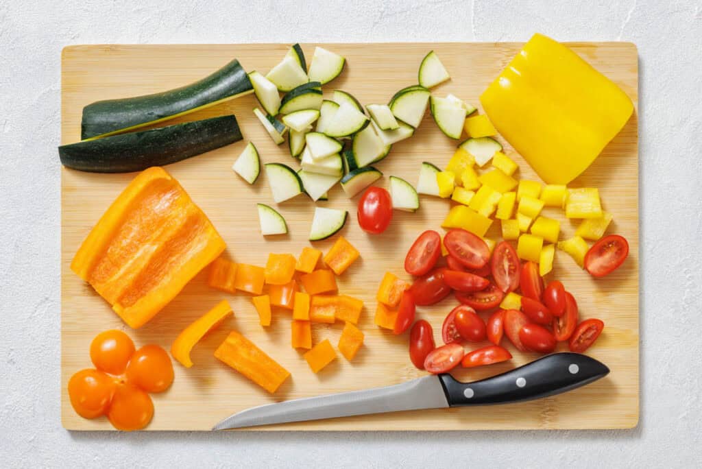 Chopped zucchini, yellow bell pepper, orange bell pepper, and halved cherry tomatoes on a wooden cutting board beside a knife.