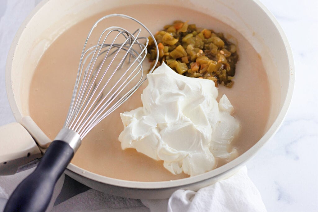 A skillet with a whisk, creamy mixture, diced green chiles, and a large dollop of sour cream on a white surface.