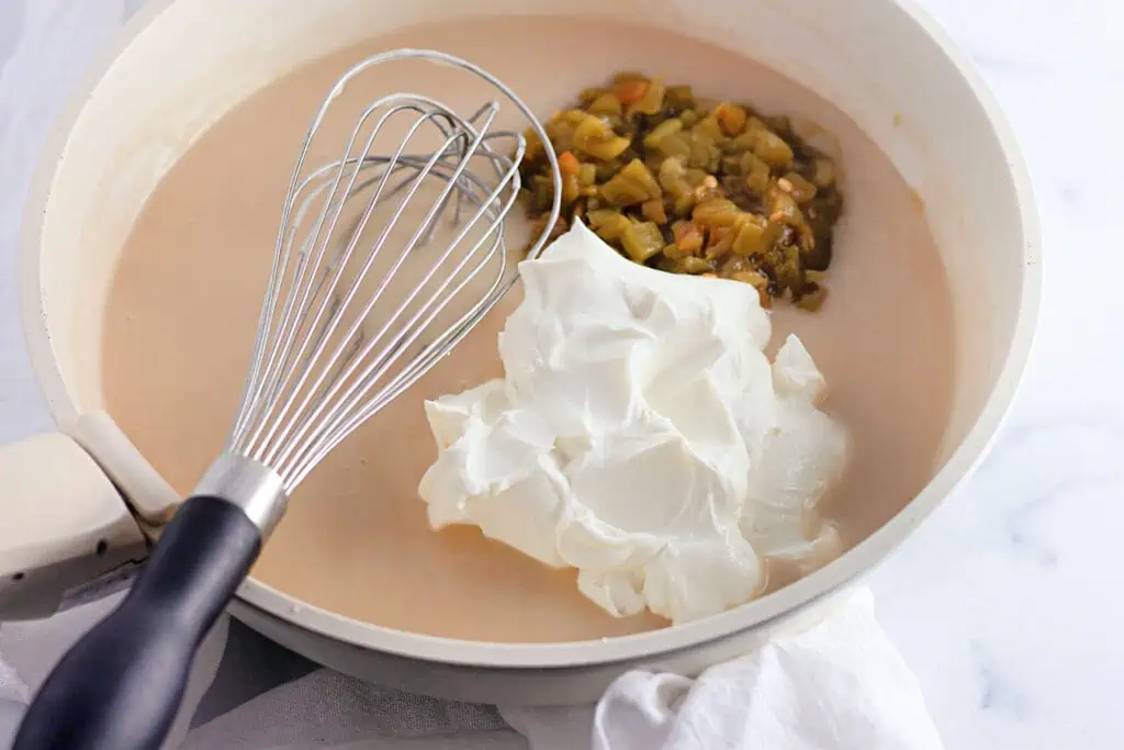 A skillet with a whisk, creamy mixture, diced green chiles, and a large dollop of sour cream on a white surface.