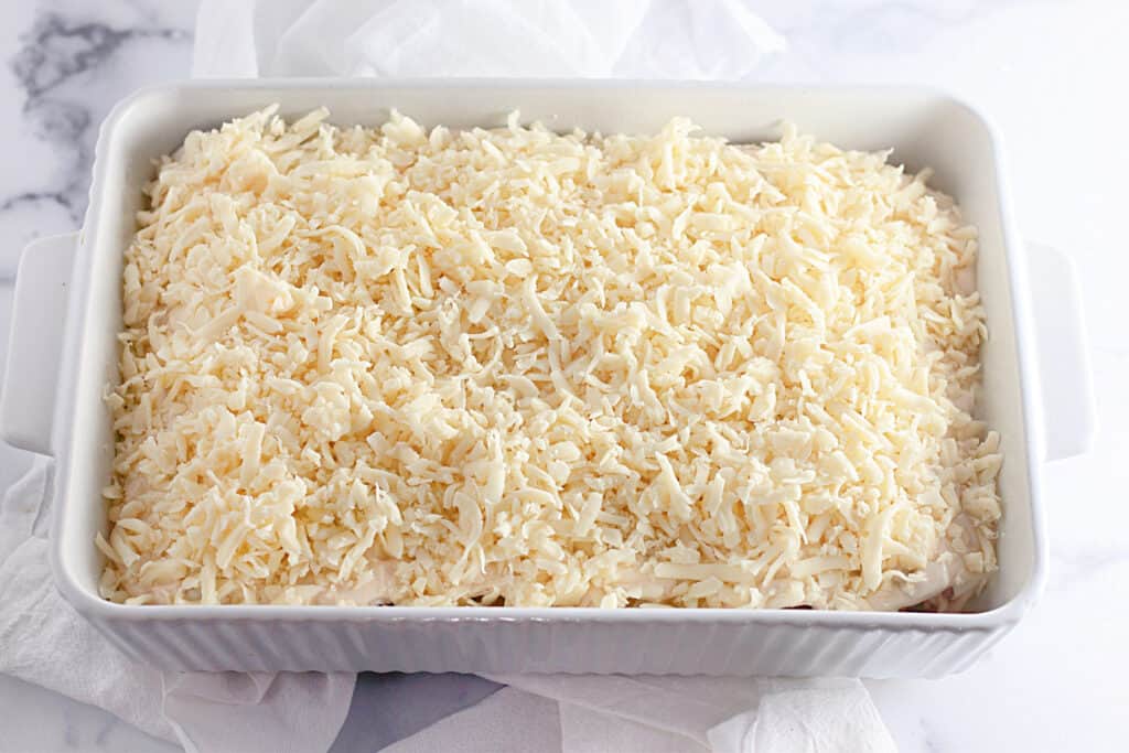 Rectangular white baking dish filled with a layer of shredded cheese on top, placed on a white cloth.