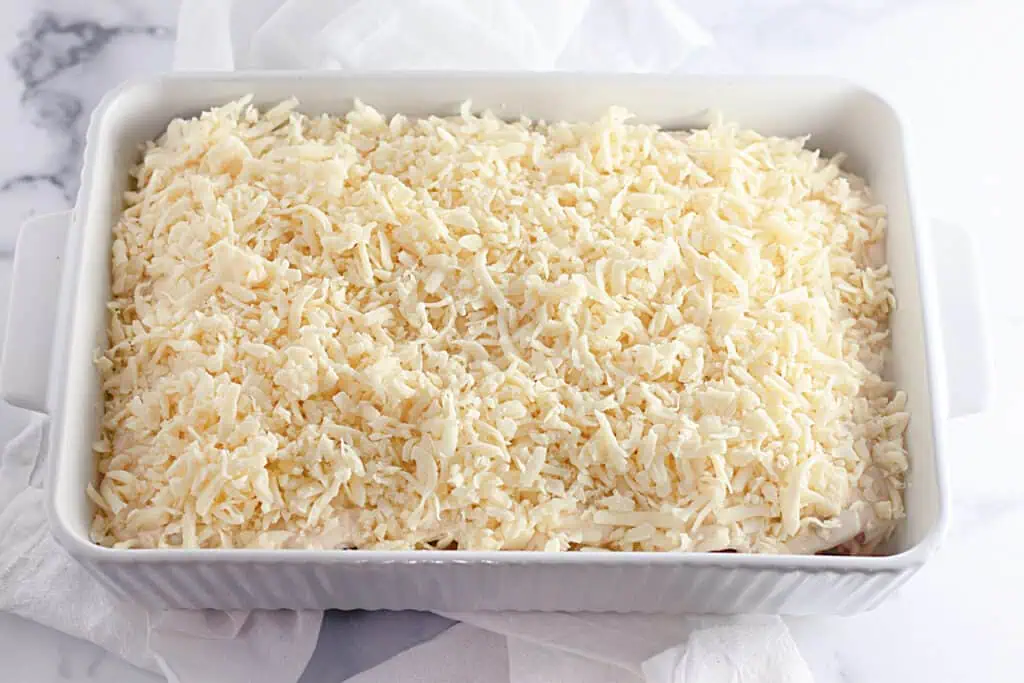 Rectangular white baking dish filled with a layer of shredded cheese on top, placed on a white cloth.