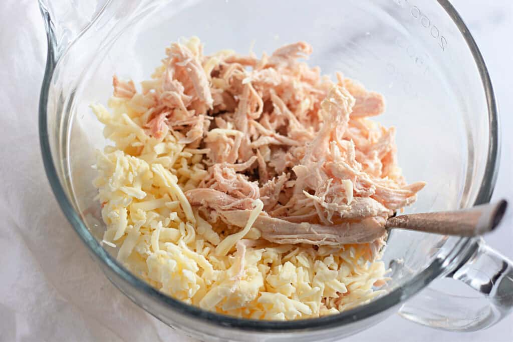 Shredded chicken and grated cheese in a glass mixing bowl with a spoon on the side.