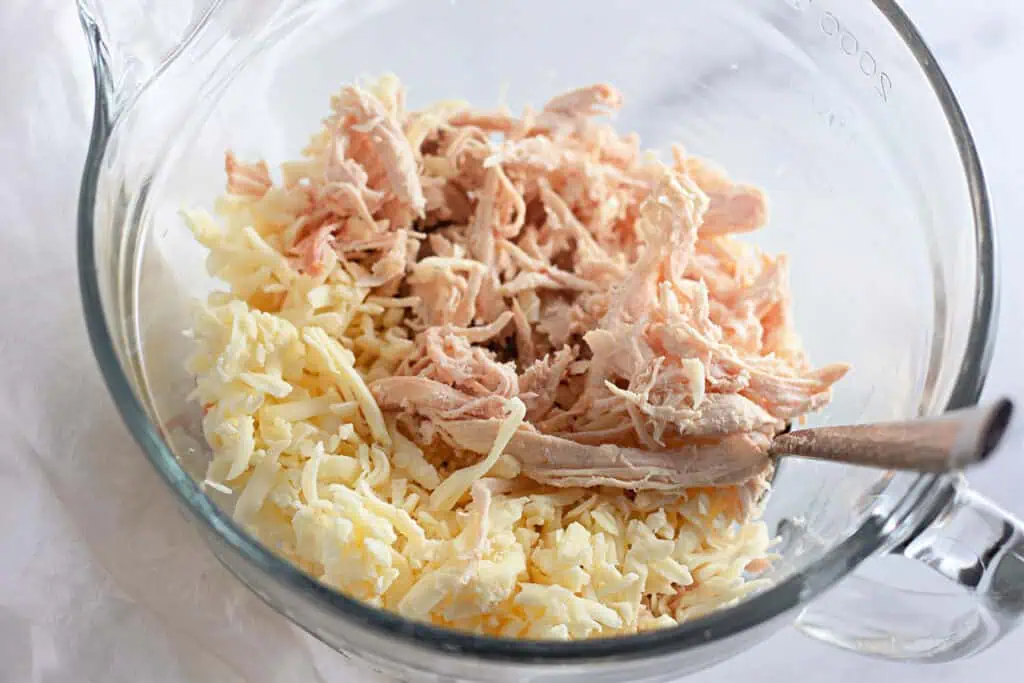 Shredded chicken and grated cheese in a glass mixing bowl with a spoon on the side.