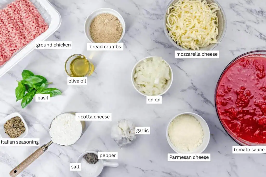 Ingredients for a recipe laid out on a marble surface, including ground chicken, cheese, tomato sauce, olive oil, breadcrumbs, onion, garlic, basil, and seasonings, each labeled.