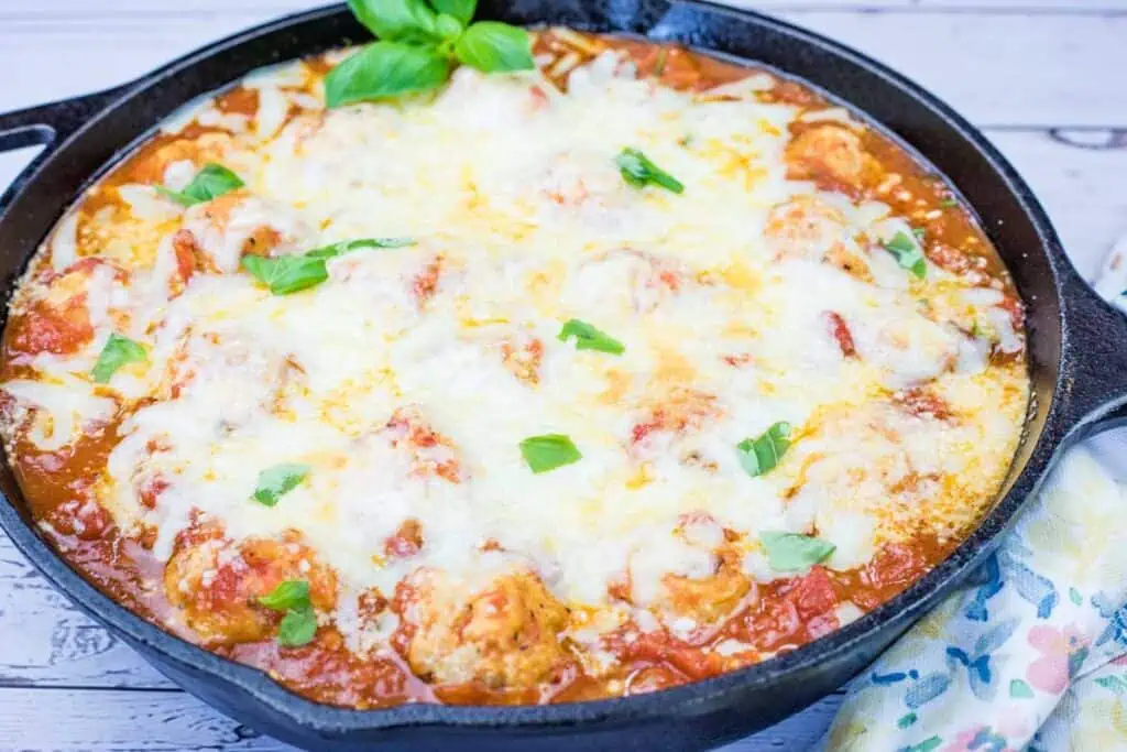 A cast iron skillet filled with baked meatballs in tomato sauce, topped with melted cheese and garnished with fresh basil leaves.
