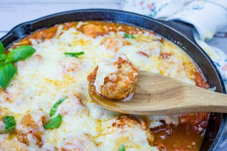 A wooden spoon holds a Chicken Parmesan Meatball over a skillet filled with meatballs.
