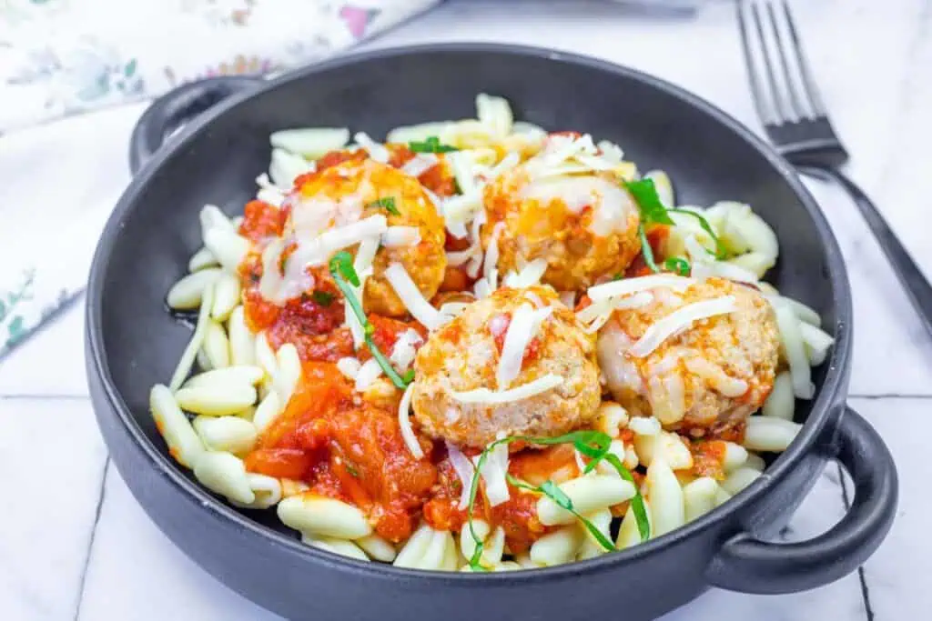 A black bowl filled with pasta, tomato sauce, and four meatballs topped with melted cheese and chopped herbs, with a fork in the background.
