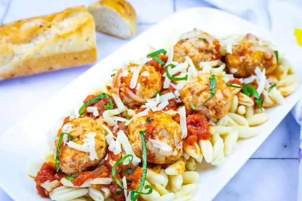 A white plate with pasta topped with meatballs, tomato sauce, shredded cheese, and fresh herbs; sliced bread is in the background.