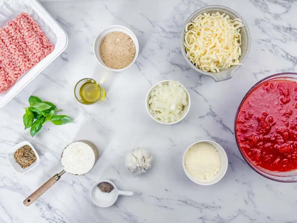 Ingredients for an Italian dish arranged on a marble surface, including ground meat, cheese, tomato sauce, garlic, onion, breadcrumbs, basil, olive oil, and seasonings.