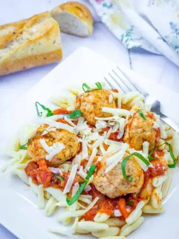 A plate of pasta topped with tomato sauce, meatballs, shredded cheese, and basil, served with a fork and a slice of bread in the background.