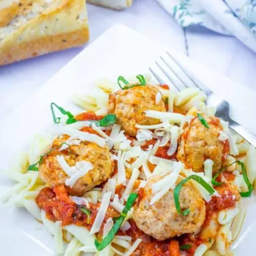 A plate of pasta topped with tomato sauce, meatballs, shredded cheese, and basil, served with a fork and a slice of bread in the background.