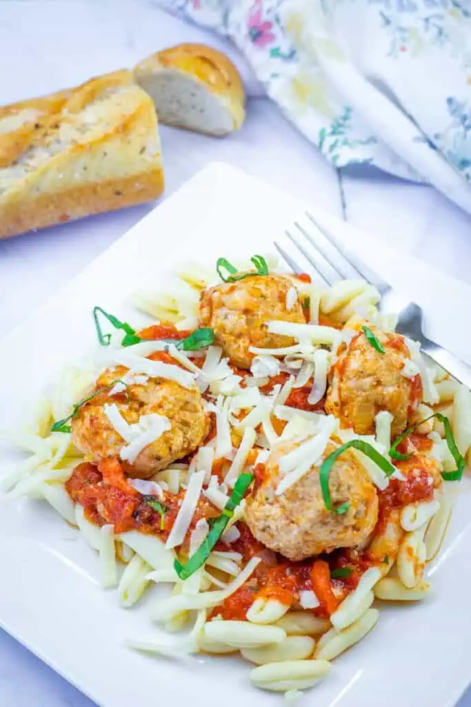 A plate of pasta topped with tomato sauce, meatballs, shredded cheese, and basil, served with a fork and a slice of bread in the background.