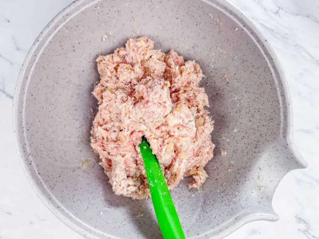 A gray mixing bowl containing raw ground meat mixture being stirred with a green spatula on a white marble surface.