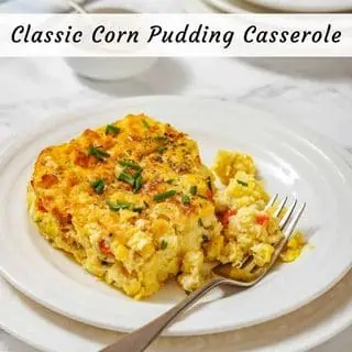 A serving of corn pudding casserole garnished with chopped chives is on a white plate, with a fork and additional plates in the background.