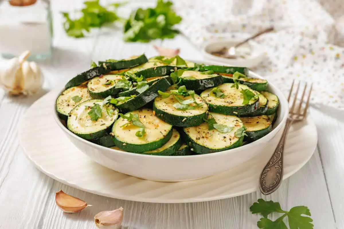 A white bowl filled with sliced Sautéed Zucchini with Garlic and Herbs, garnished with chopped herbs, is placed on a white plate with a fork nearby.
