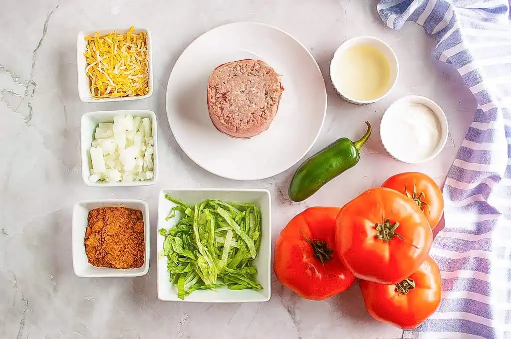 Overhead shot of labeled ingredients for taco stuffed tomatoes, including ground beef, tomatoes, cheese, onion, lettuce, taco seasoning, jalapeño, olive oil, and sour cream.
