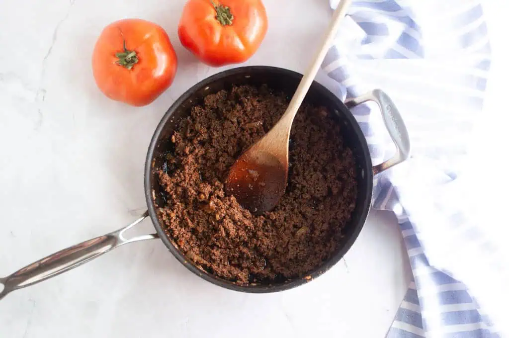 Overhead shot of seasoned ground meat being stirred in a black skillet with a wooden spoon.