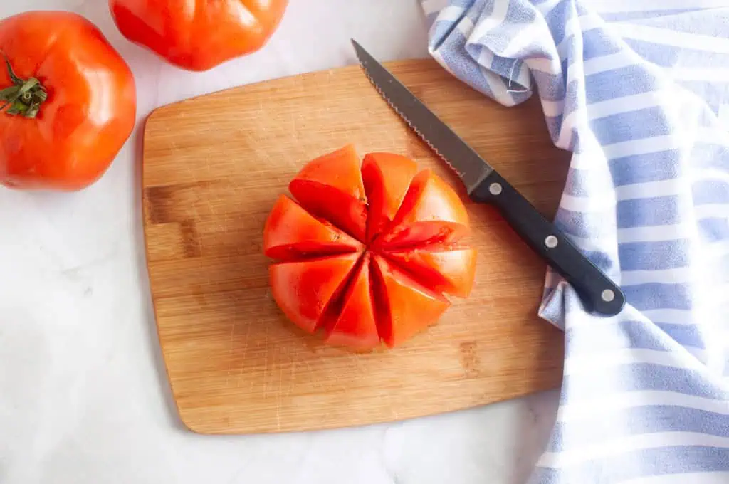 Overhead shot of a tomato partially sliced into wedges on a wooden cutting board with a knife.