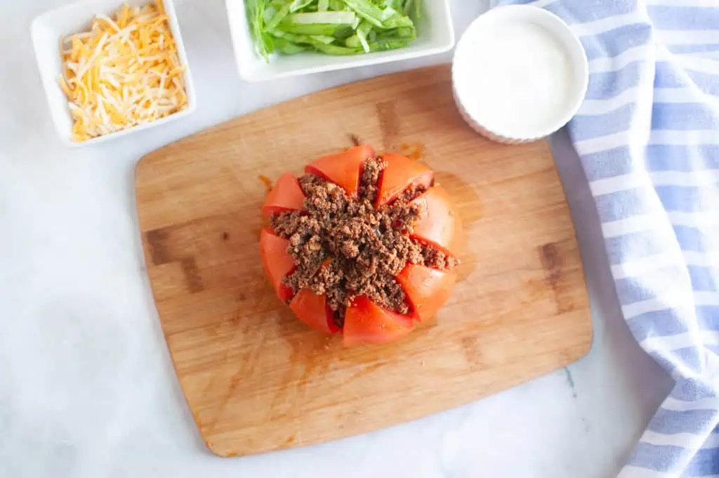 Overhead shot of a tomato being filled with seasoned ground lamb on a wooden cutting board.