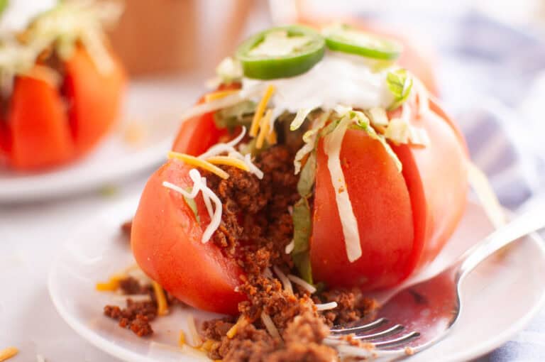 A sliced tomato stuffed with seasoned ground meat, shredded cheese, lettuce, sour cream, and jalapeño slices is served on a white plate with a fork.