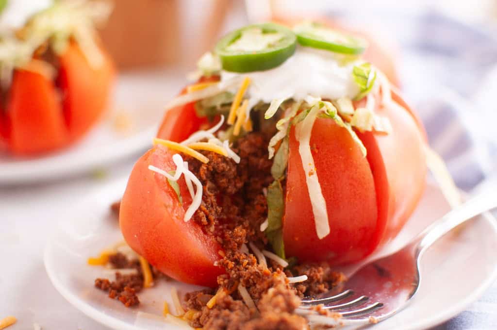A sliced tomato stuffed with seasoned ground meat, shredded cheese, lettuce, sour cream, and jalapeño slices is served on a white plate with a fork.