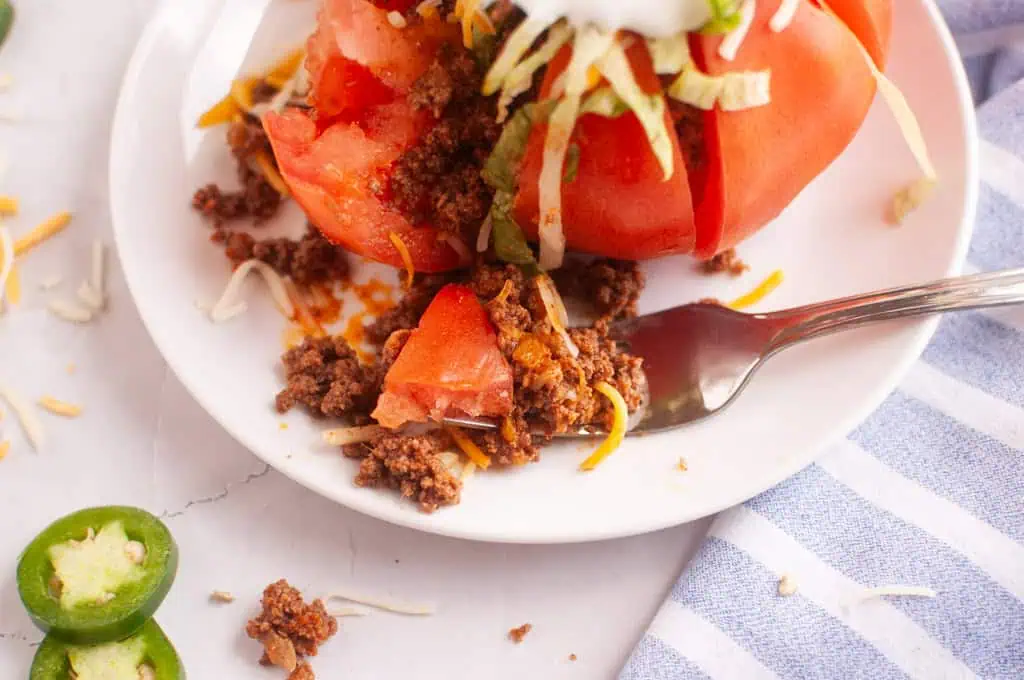 A fork takes a bite out of a taco stuffed tomato on a white plate.