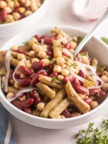A bowl of three-bean salad with kidney beans, chickpeas, onions, and herbs sits on a table next to a blue striped napkin and fresh parsley.