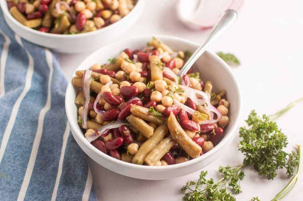 A bowl of three-bean salad with kidney beans, chickpeas, onions, and herbs sits on a table next to a blue striped napkin and fresh parsley.