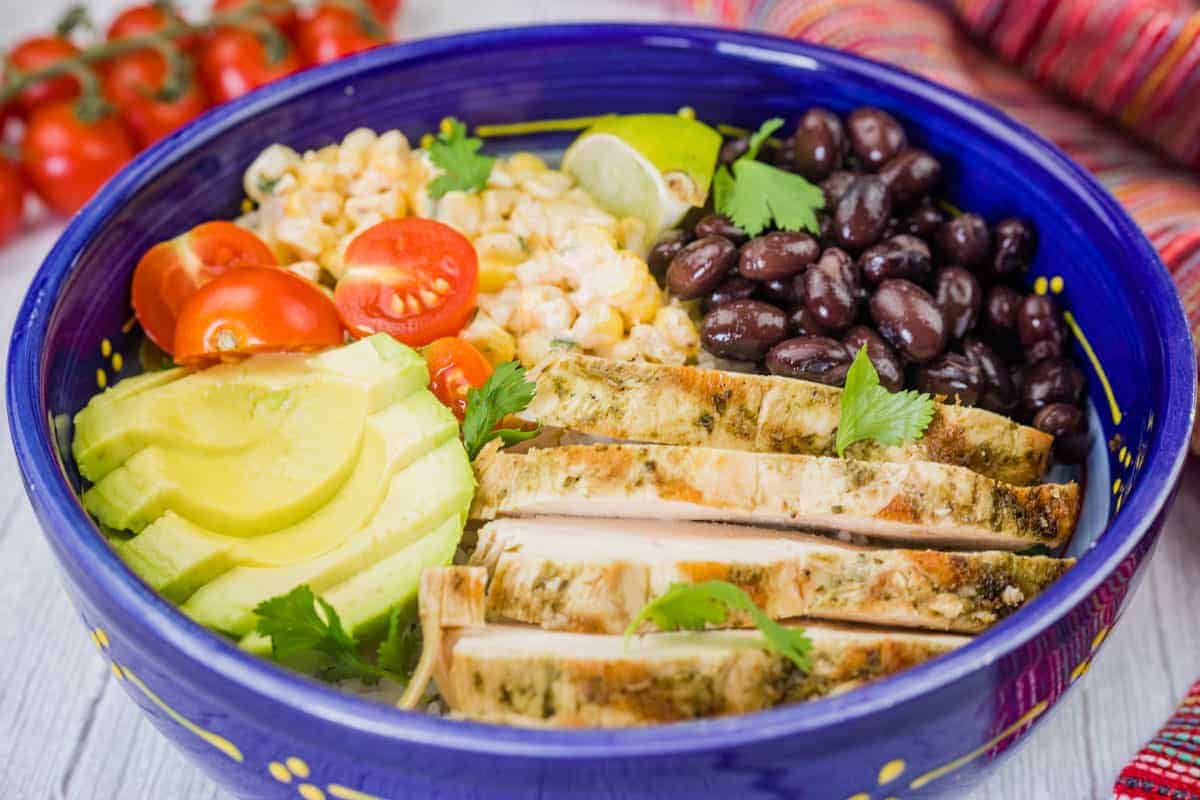 A blue bowl filled with street corn chicken rice bowl, featuring grilled chicken, corn, tomatoes, avocado, and black beans.