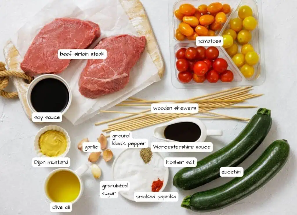 Ingredients for steak skewers arranged on a white surface, including beef sirloin, tomatoes, zucchini, sauces, spices, garlic, olive oil, and wooden skewers.