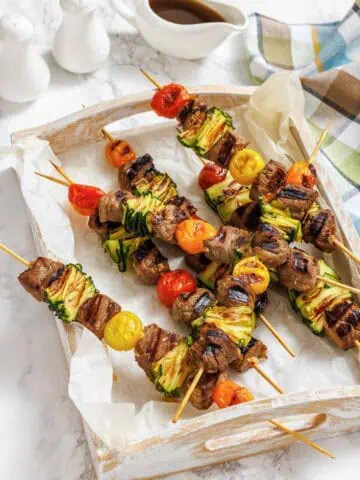Four grilled meat and vegetable skewers with zucchini and cherry tomatoes on a parchment-lined wooden tray, with a sauce boat and salt and pepper shakers in the background.
