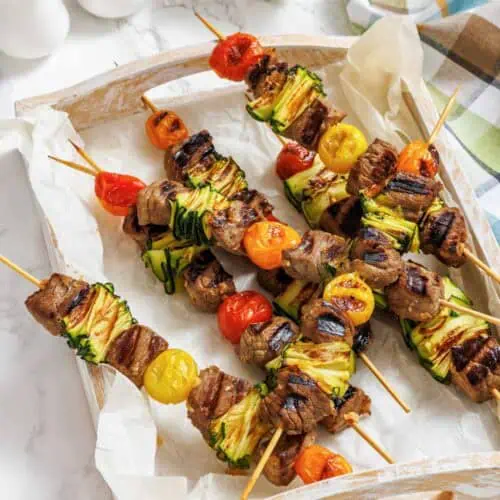 Four grilled meat and vegetable skewers with zucchini and cherry tomatoes on a parchment-lined wooden tray, with a sauce boat and salt and pepper shakers in the background.