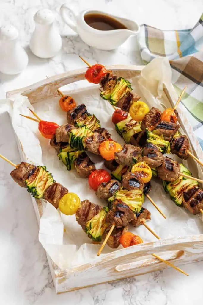 Four grilled meat and vegetable skewers with zucchini and cherry tomatoes on a parchment-lined wooden tray, with a sauce boat and salt and pepper shakers in the background.