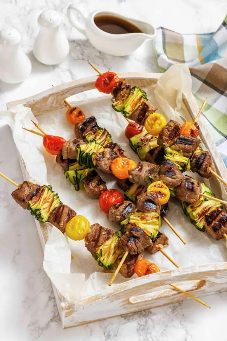 Four grilled meat and vegetable skewers with zucchini and cherry tomatoes on a parchment-lined wooden tray, with a sauce boat and salt and pepper shakers in the background.