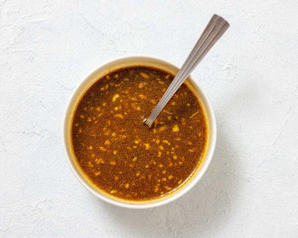 A white bowl filled with a brown liquid sauce with visible minced garlic, and a metal spoon resting inside, on a light textured surface.