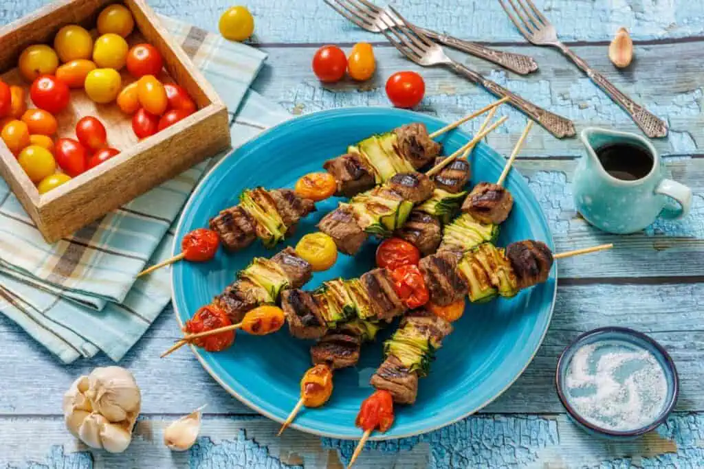 A blue plate holds grilled meat and vegetable skewers with cherry tomatoes, zucchini, and mushrooms; a box of tomatoes and small dishes are nearby on a rustic blue wooden table.