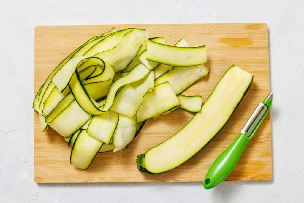 Zucchini sliced into thin ribbons with a green vegetable peeler on a wooden cutting board.