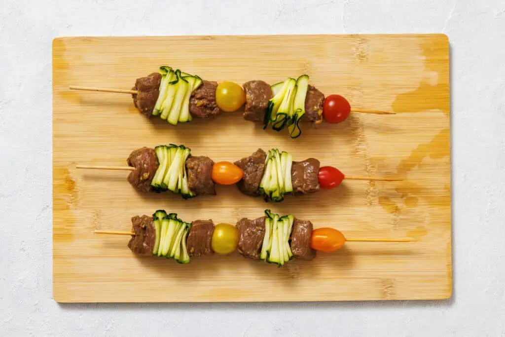 Four uncooked skewers with pieces of beef, zucchini ribbons, and cherry tomatoes on a wooden cutting board.